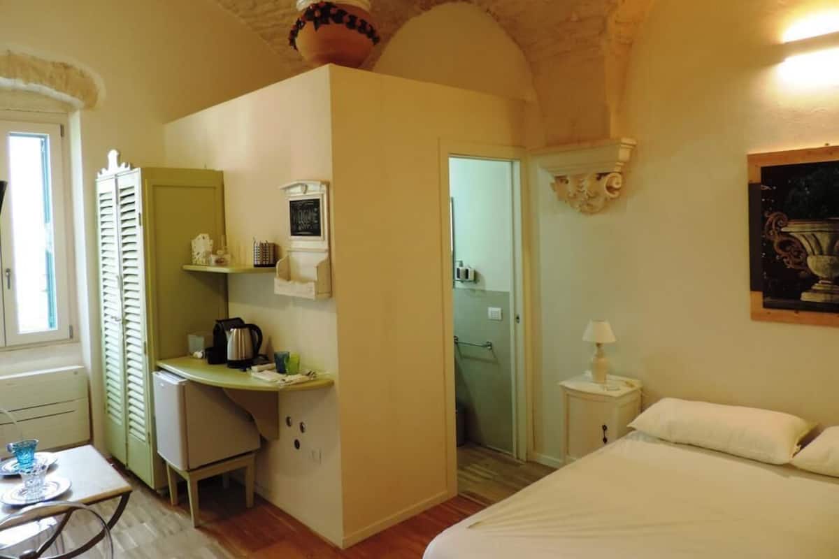 Une petite chambre comprenant un lit, un petit bureau avec une bouilloire, une chaise et une fenêtre.