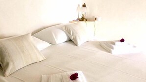 Ein ordentlich bezogenes Bett mit weißen Leinentüchern, ein gefaltetes Handtuch und eine Rose auf dem Bett. Ein Nachttisch mit Lampe und ein Gemälde an der Wand über dem Bett.