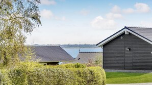 En rad mörka trähus med tegeltak, en gräsyta och en vattenyta i bakgrunden.
