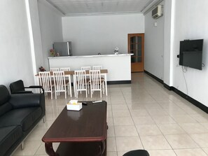 A living room with a black sofa, a wooden coffee table, and a dining area with white chairs and a small kitchen area with a refrigerator and a TV.