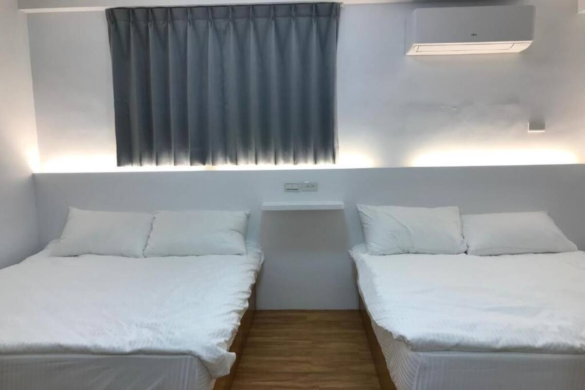 A room with two single beds, a small table, a wall-mounted air conditioner, and a window with curtains.