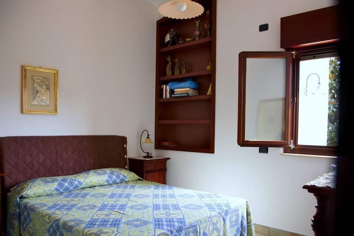 Un dormitorio con cama, una mesita de noche, una estantería con objetos decorativos, un cuadro en la pared y una ventana con vistas a zonas verdes.