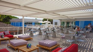A covered outdoor seating area with tables and chairs, a pool, and a building in the background.