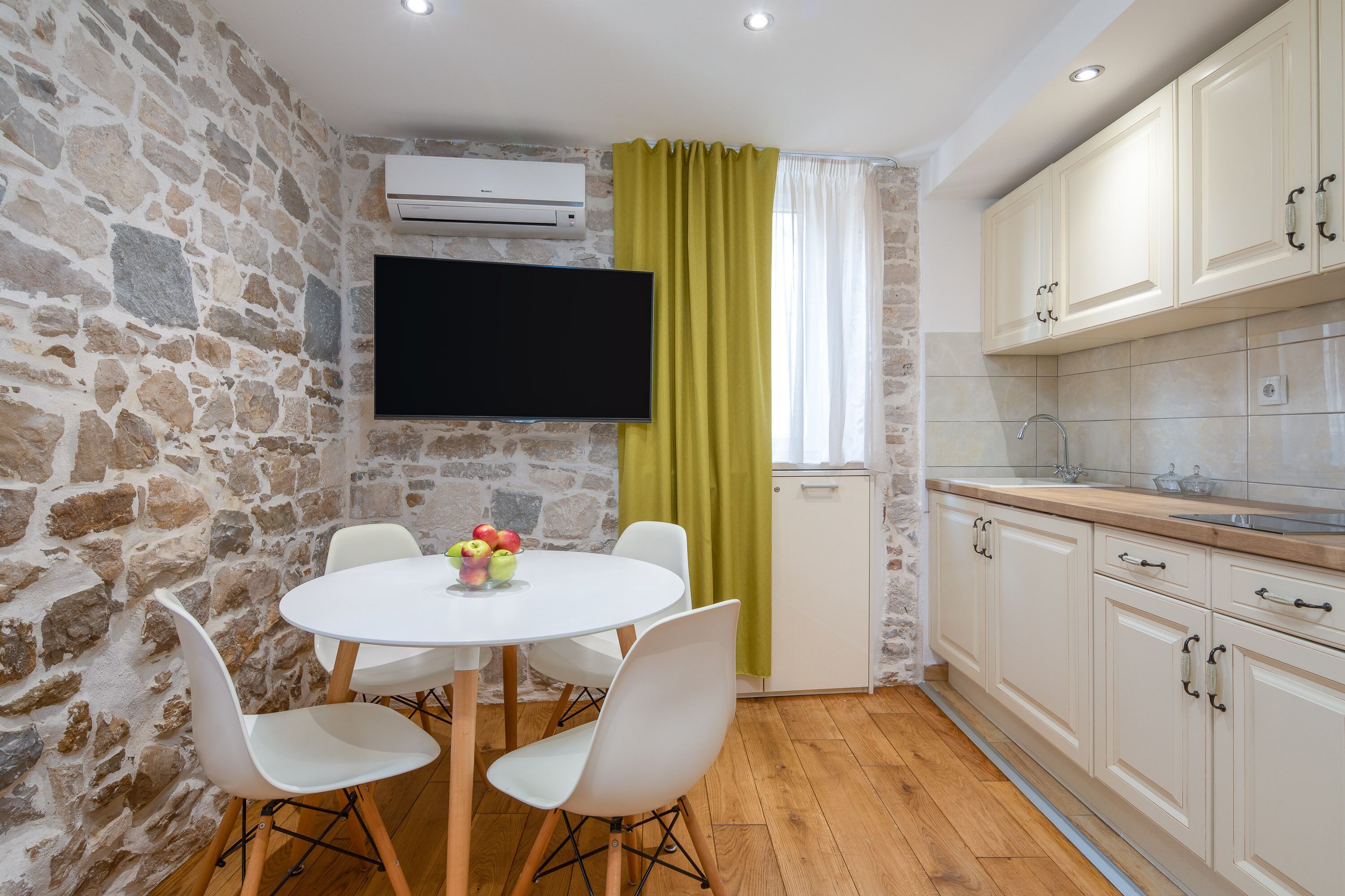 A compact kitchen with a stone wall, a flat-screen TV, a dining table with chairs, and a window with curtains.