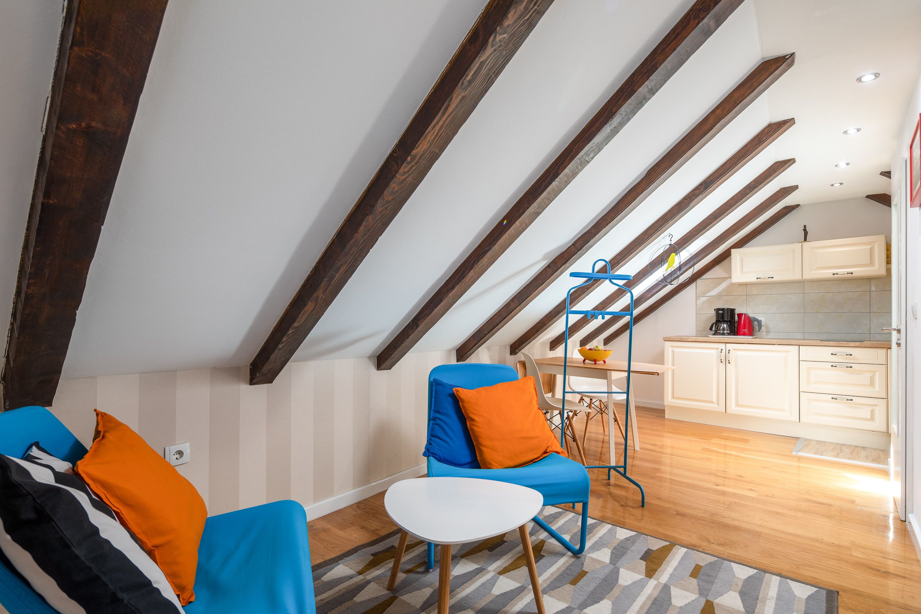 Una habitación moderna estilo loft con vigas de madera a la vista, un sillón azul con cojines naranjas, una mesita blanca y una zona de cocina con armarios blancos y una cafetera.
