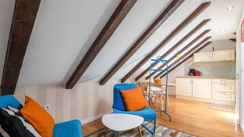Una habitación moderna estilo loft con vigas de madera a la vista, un sillón azul con cojines naranjas, una mesita blanca y una zona de cocina con armarios blancos y una cafetera.