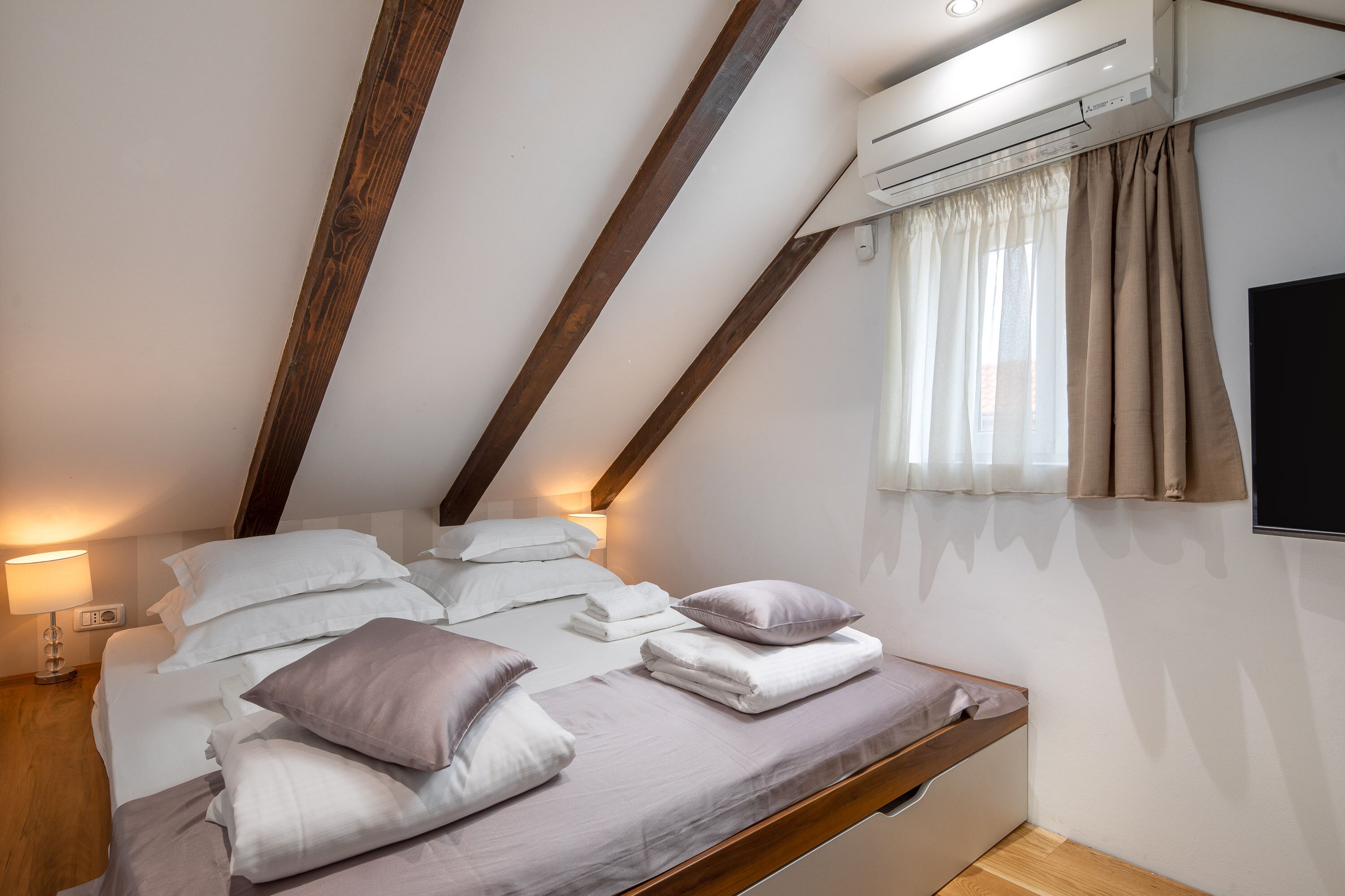 A bedroom with a sloped ceiling, a bed with white and purple bedding, a bedside lamp, and an air conditioning unit.