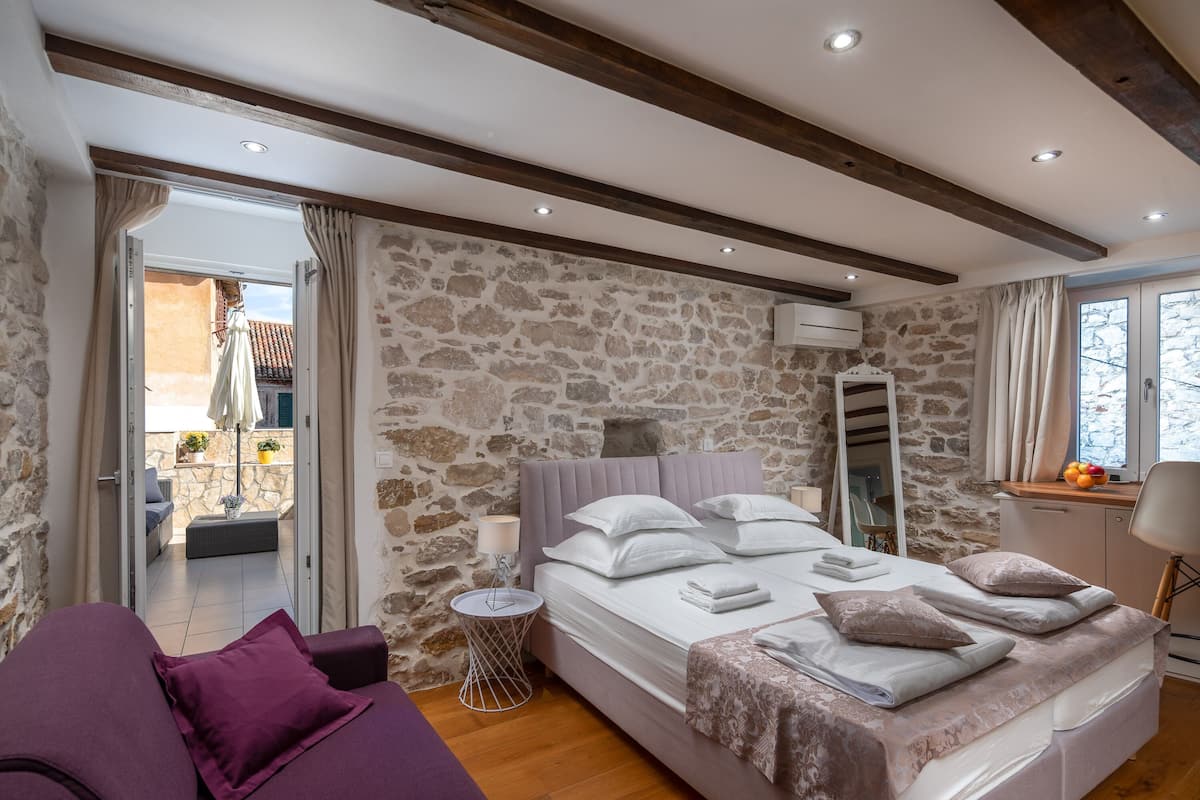 A bedroom with a stone wall, a bed with pillows and a blanket, a small round table with a lamp, and a purple sofa.