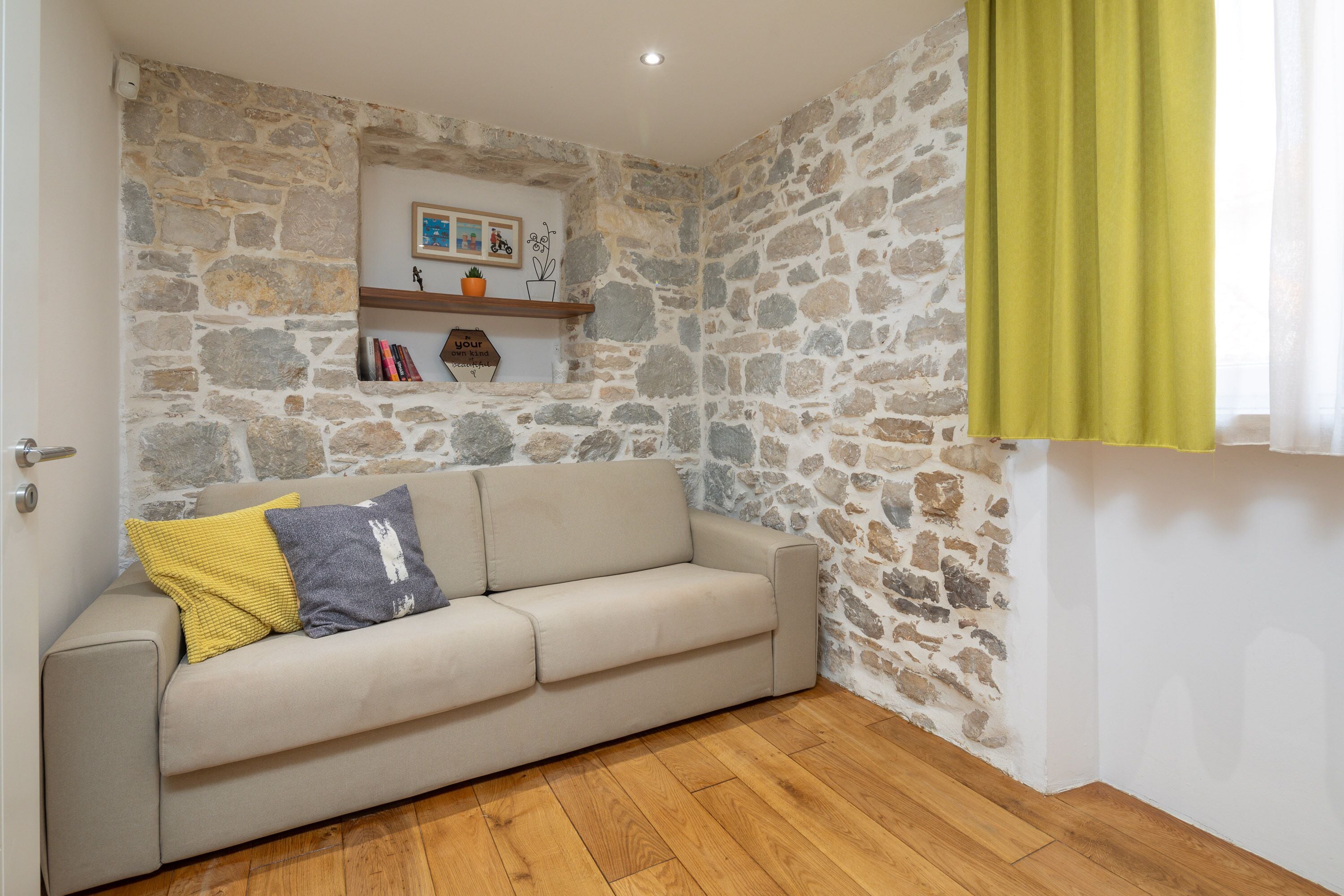Ein Zimmer mit einer Steinwand, einem beige farbenen Sofa mit Kissen, Holzboden und einem grünen Vorhang.