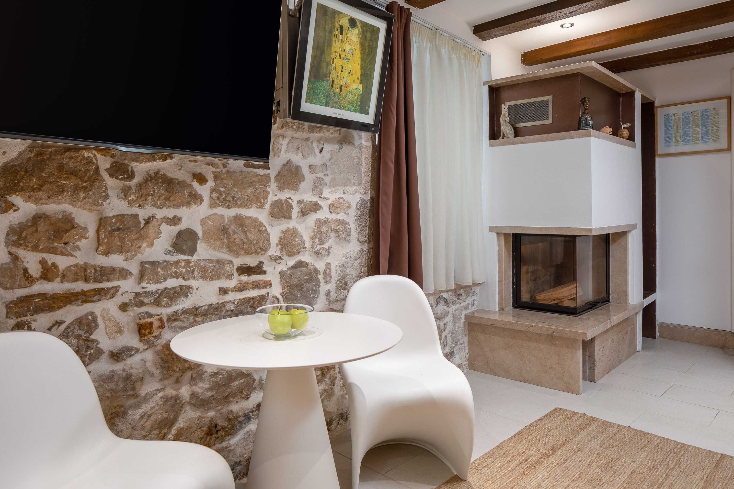 A modern living room with a stone wall, a fireplace, a white round table with a bowl of fruit, and two white chairs.
