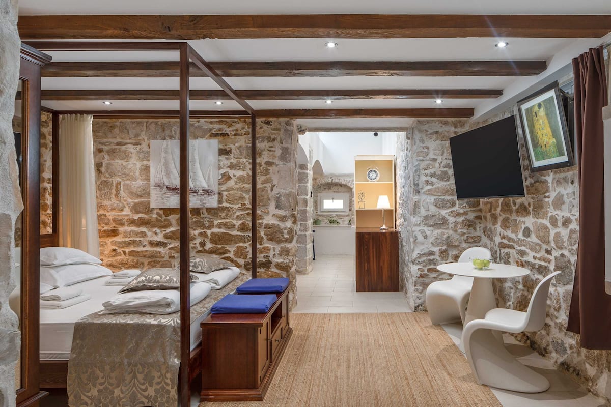 A room with a stone wall, a bed with a canopy, a small table with chairs, and a television mounted on the wall.