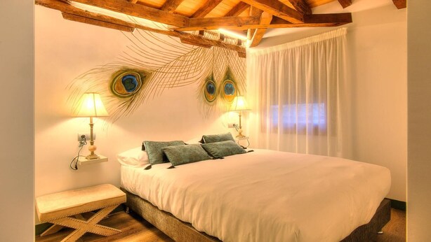 A bedroom with a wooden ceiling, a bed with white bedding, two wall-mounted lamps, and a peacock feather wall decoration. - SYNAGOGUE 6 DELUXE WITH PRIVATE TERRACE (Toledo)
