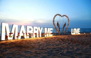 A beach setting with large illuminated letters spelling "MARRY ME LOVE" and a heart-shaped decoration made of branches and flowers.