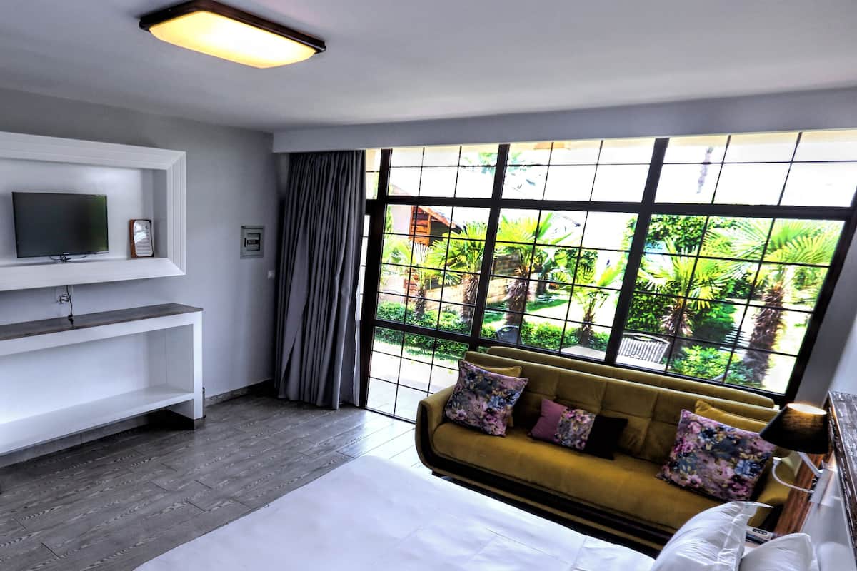 A modern hotel room with a large bed, a sofa with floral cushions, a flat-screen TV mounted on the wall, and a view of greenery through large windows.