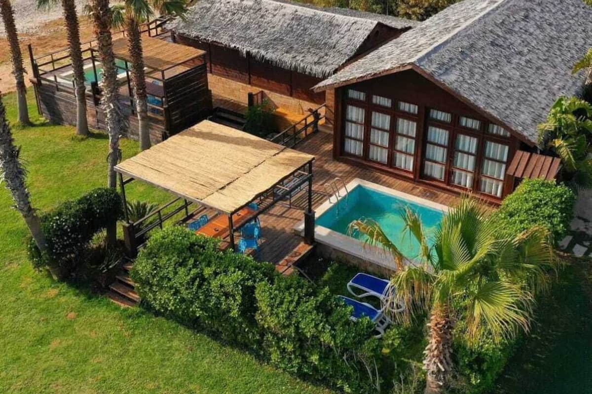 A bungalow with a pool, surrounded by lush greenery and palm trees, overlooking a beach.