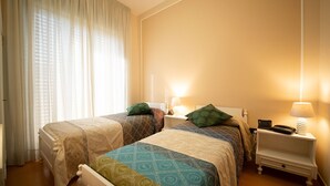 A hotel room with two beds, each with patterned bedding and pillows, a nightstand with a lamp and phone, and a window with sheer curtains allowing natural light. - Hotel Ristorante Venezuela (Montesano sulla Marcellana)