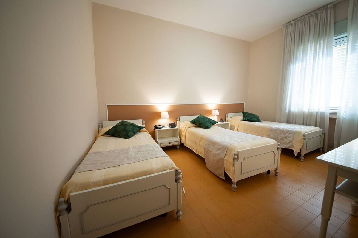 A hotel room with three single beds, each with a bedside table and lamp. The room has a wooden floor and a large window with curtains.