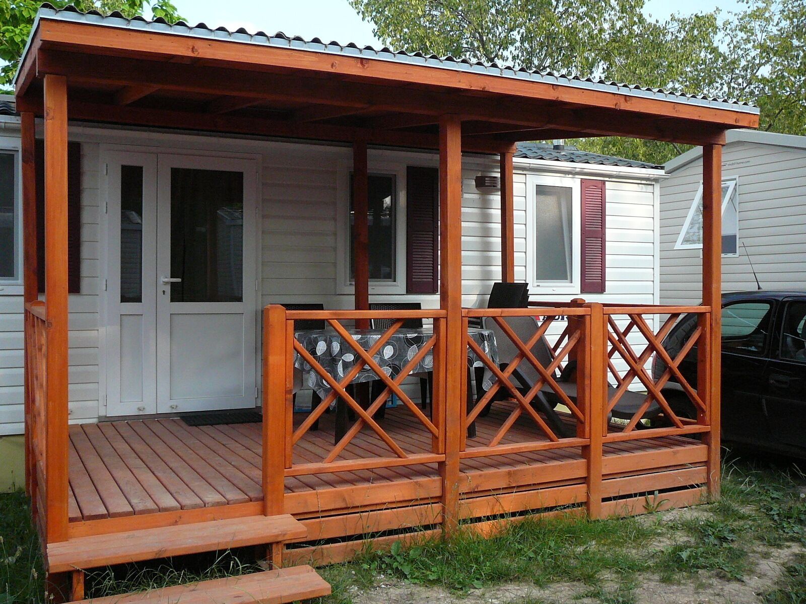 Une terrasse en bois avec une rambarde, un porche couvert et une voiture garée à proximité.