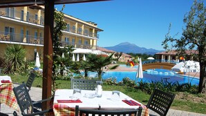 A poolside dining area with tables set for two, surrounded by greenery and a view of a mountain.