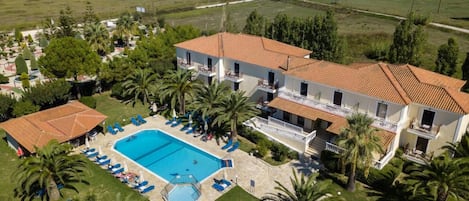 Un edificio de dos pisos con piscina, rodeado de palmeras y un césped cuidado.