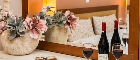 Habitación de hotel con cama, mesita de noche con dos botellas de vino y copas, un espejo y una planta decorativa.
