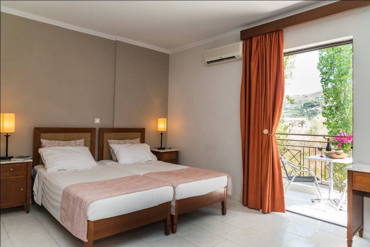 Habitación de hotel con cama doble, dos mesitas de noche con lámparas, un balcón con mesa y sillas, y un aire acondicionado.