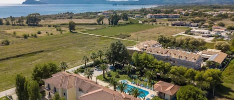 Un complejo turístico en la costa con varias edificaciones, piscina y vista al mar y a las montañas.