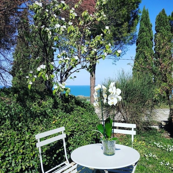A round table with a vase of white flowers, two white chairs, and a view of the sea and trees.