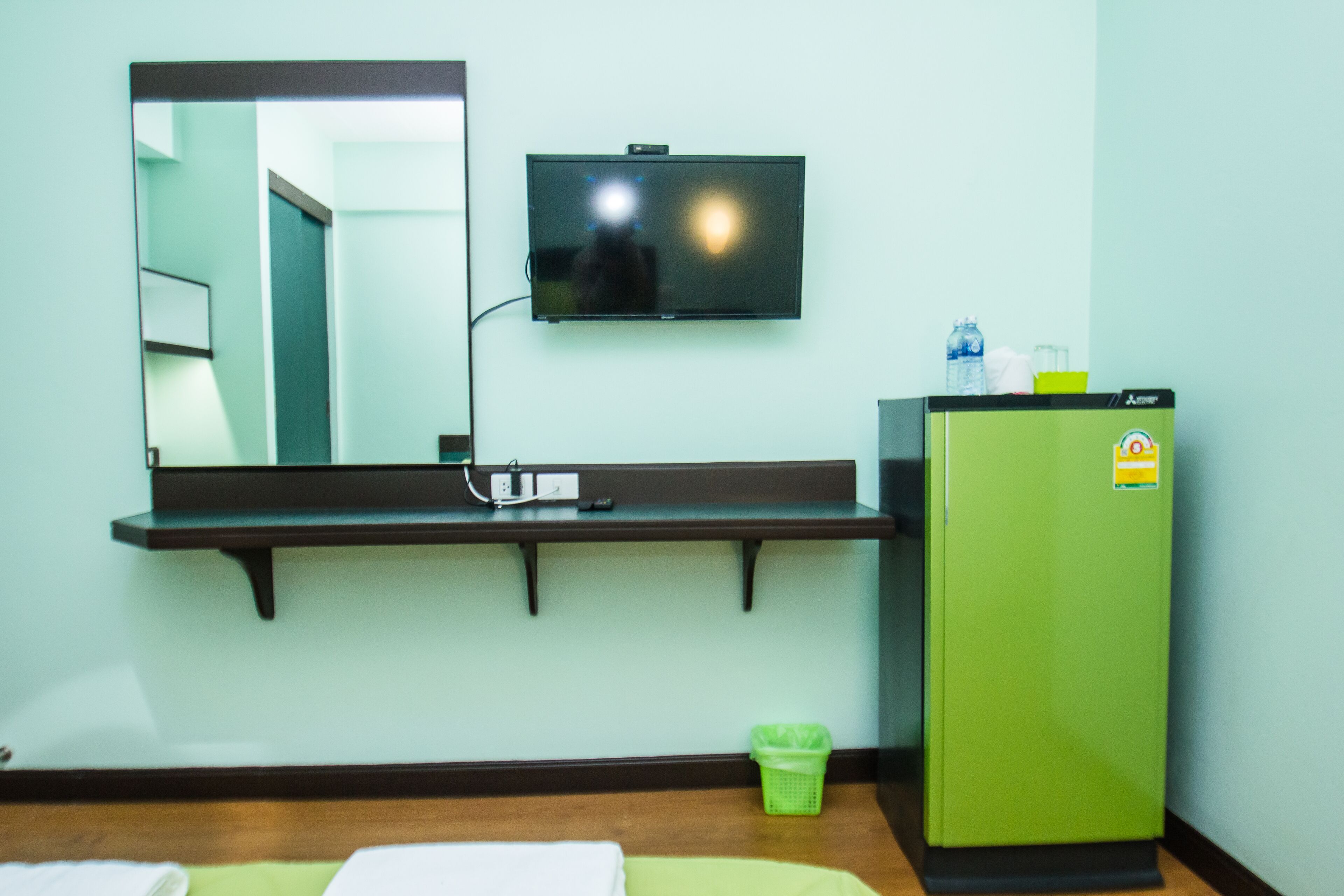 A hotel room with a bed, a green minibar, a television mounted on the wall, and a mirror above the minibar.