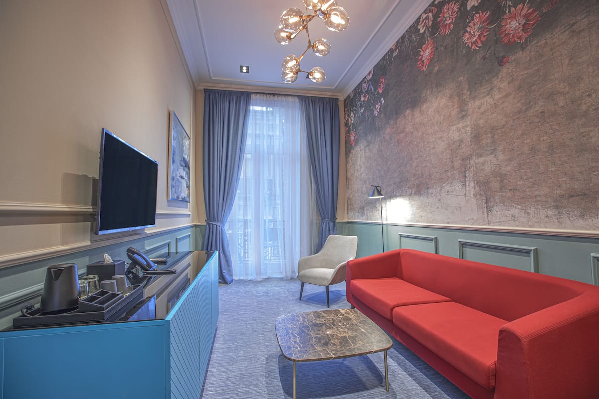 A hotel room with a red sofa, a coffee table, a TV, a desk with a telephone and a lamp, and a large floral mural on the wall.