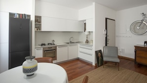 Une cuisine compacte avec des meubles blancs, un réfrigérateur, un évier et une cuisinière. On y trouve une table à manger avec un vase et une chaise. Un vélo est accroché au mur.