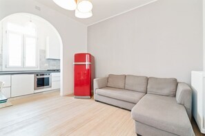 Une cuisine moderne avec des meubles blancs, un appareil électroménager rouge et un canapé beige dans une pièce lumineuse dotée d’un plancher en bois.