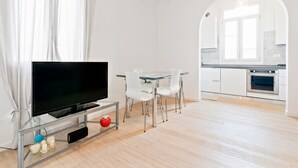 Un soggiorno moderno con TV a schermo piatto, tavolo da pranzo in vetro, sedie bianche e angolo cottura con mobili bianchi e forno.