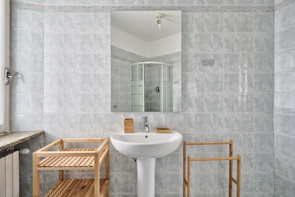 A bathroom with a white pedestal sink, wooden shelves, and a glass shower enclosure. - GuestHost - Comfortable attic apartment with a large furnished terrace is perfect for 3 people, it is located a few meters from the Carducci metro station. We are also a stone's throw from the hospitals (CTO, Molinette, Cellini) and the Parco del Valenti (Torino)