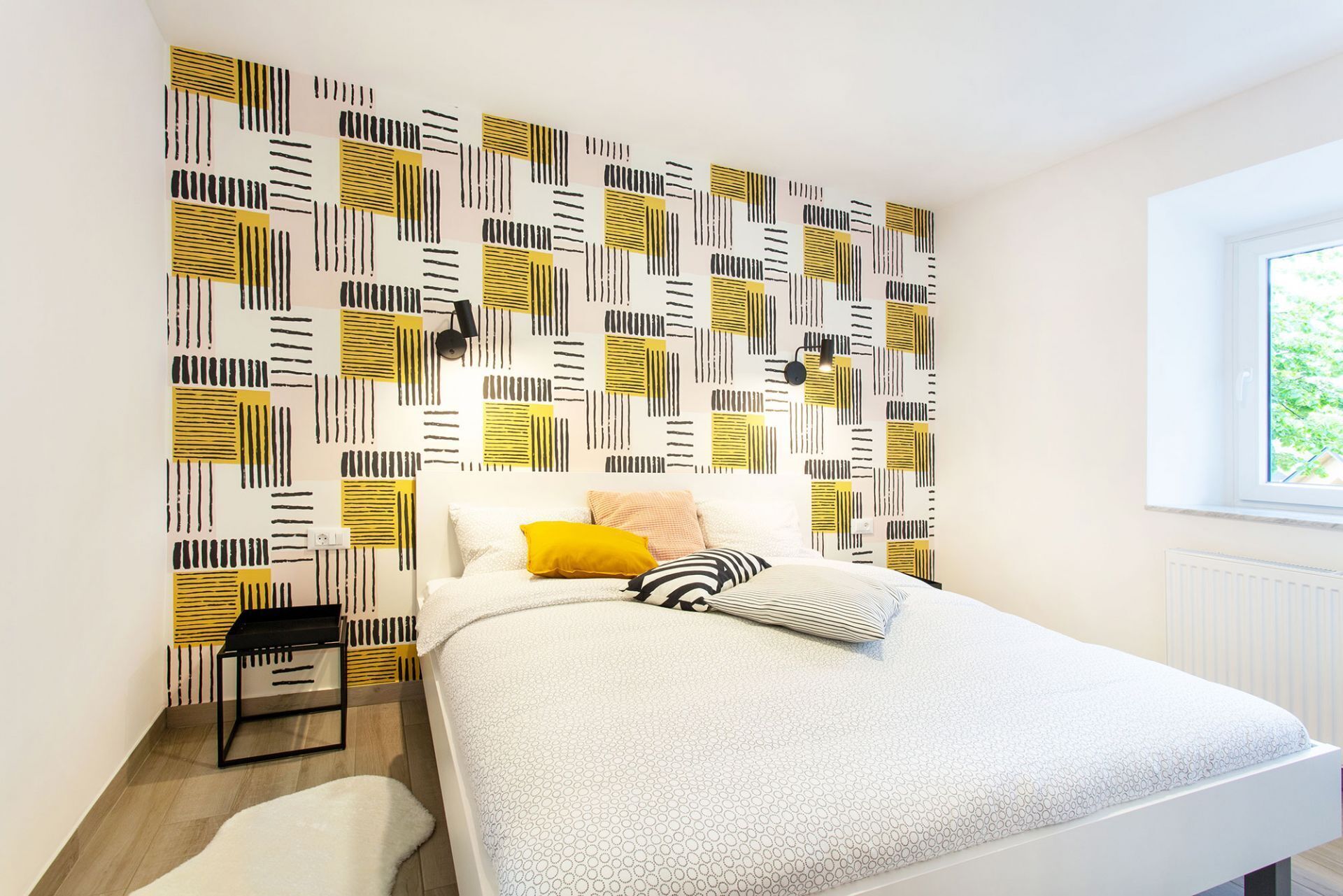 Un dormitorio con papel tapiz estampado, una cama con ropa blanca, una ventana con vistas y una mesita de noche pequeña.