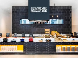 Interior de una cafetería moderna con pared de ladrillo negro, un logotipo sobre la barra y diversos alimentos exhibidos.