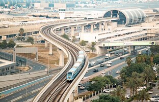 A train on elevated tracks curving around a modern building with a curved roof, passing under a bridge labeled "Cartlow."