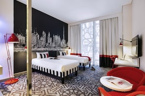 Una habitación de hotel con una cama grande, dos mesitas de noche, un sillón rojo, una pequeña mesa redonda y un mural de paisaje urbano en la pared.