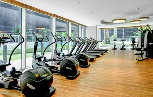 A gym with multiple treadmills lined up against large windows, a wooden floor, and various exercise machines.
