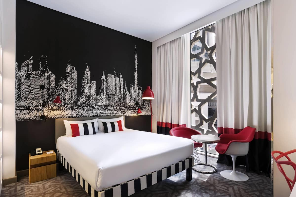 A modern hotel room with a large bed, a cityscape mural, a desk with a lamp, and red chairs.
