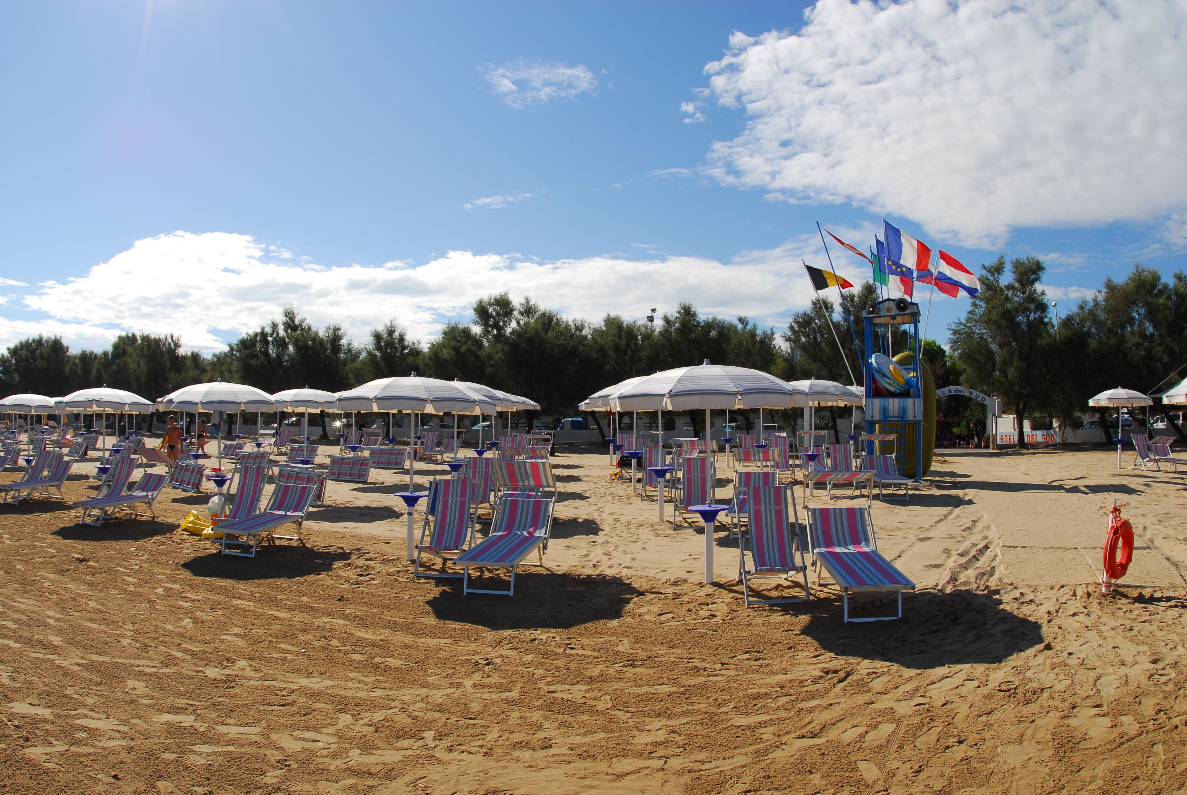 Ein Strand mit Reihen von gestreiften Liegestühlen und Sonnenschirmen, einem Rettungsring und Flaggen.