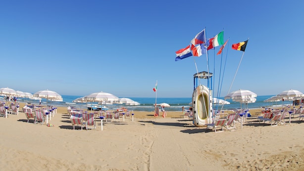 A beach with rows of beach chairs and umbrellas, flags, and a lifeguard tower. - Victoria Mobilehome Stella del Sud Camping Villaggio (Rodi Garganico)