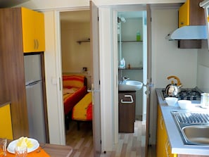 Eine kleine Küche mit Kühlschrank, Spüle und Herd. Im angrenzenden Raum ist ein Bett mit einer rot-gelb gestreiften Decke zu sehen.