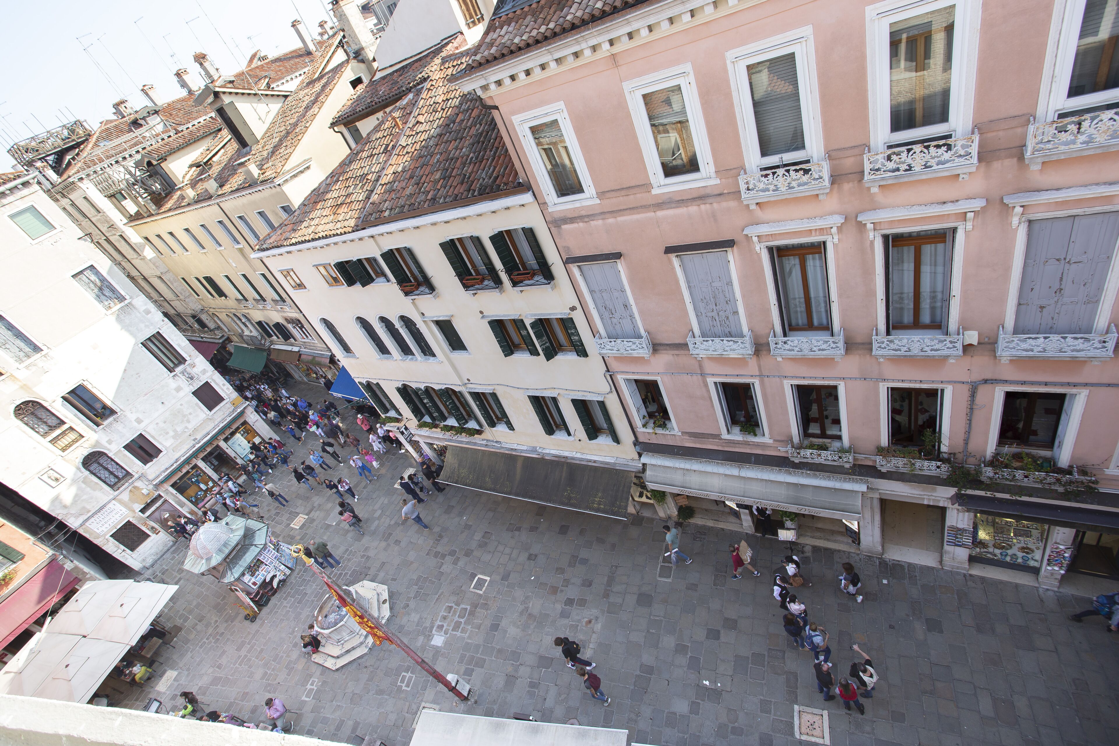 A crowded square with historic buildings, including a pink building with balconies and a white building with a shop at the corner.