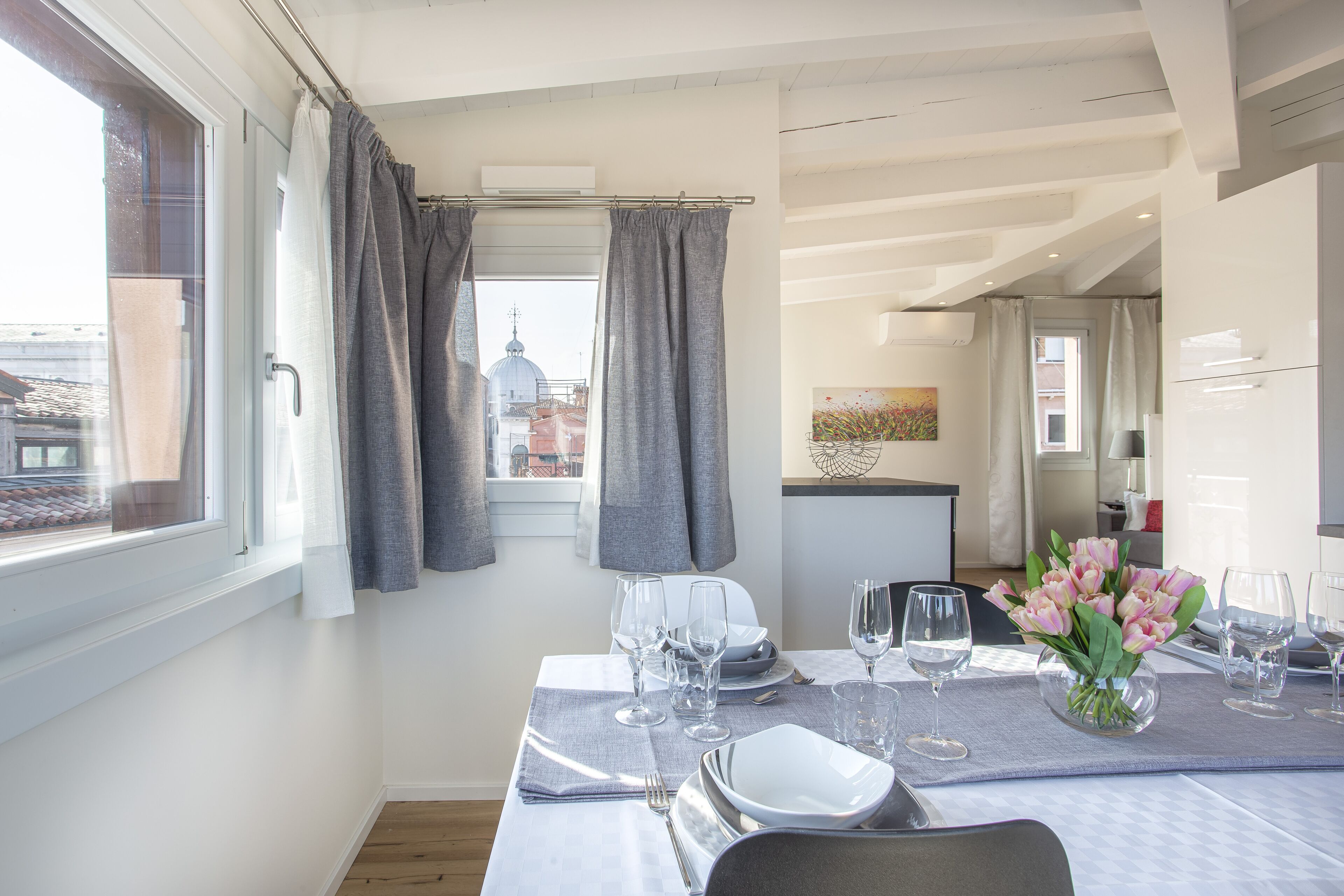 A dining area with a table set for four, a vase of flowers, and a view of buildings through the window.