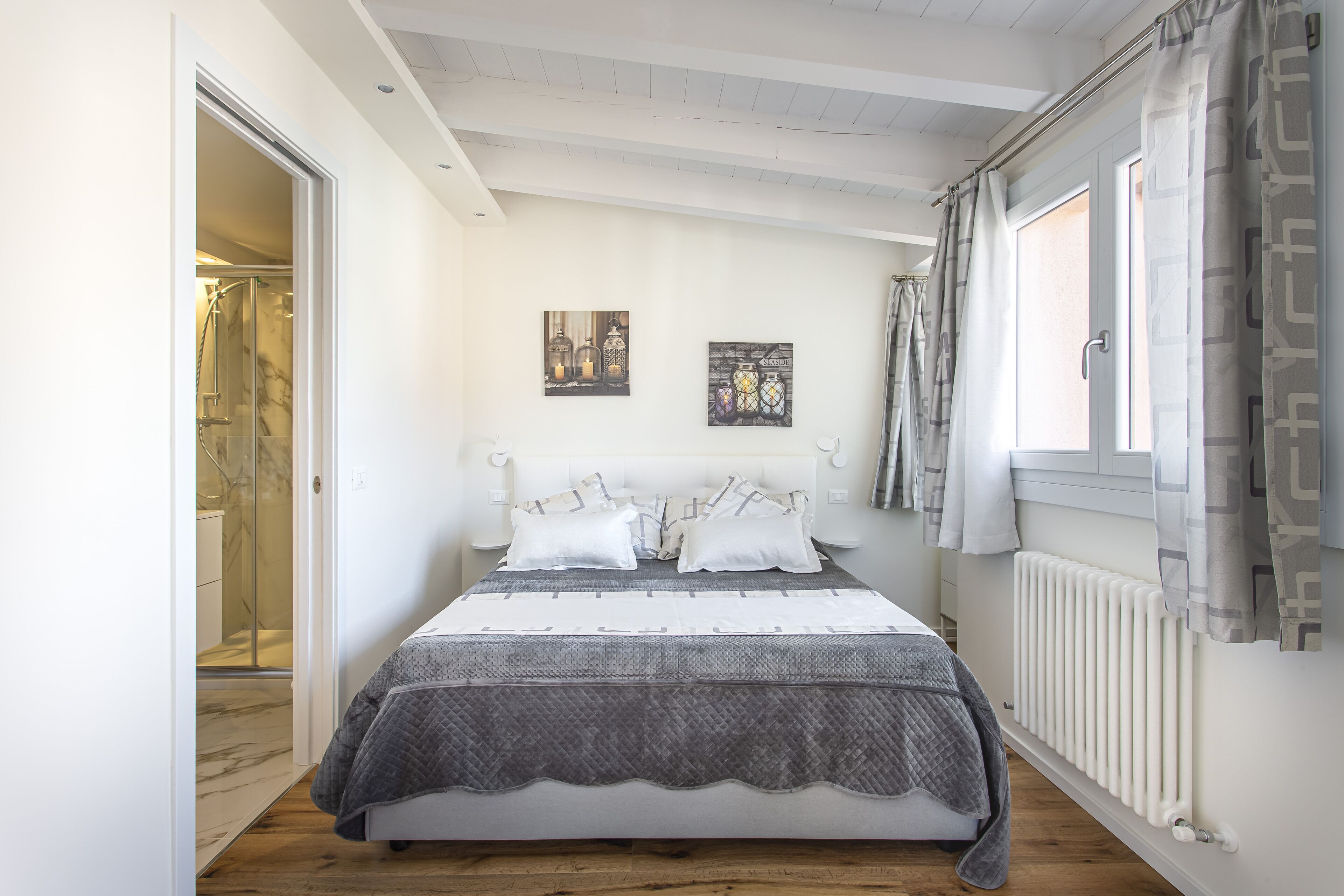 A bedroom with a bed, a window with curtains, a radiator, and a door leading to a bathroom.