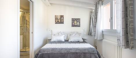 A bedroom with a bed, a window with curtains, a radiator, and a door leading to a bathroom.