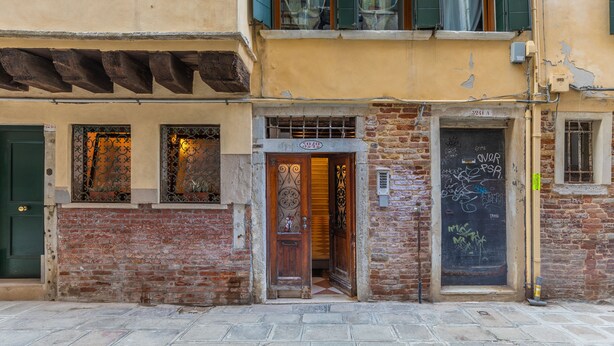 A building with a wooden door, a window with a metal grill, and a brick facade. - Venice Rio dei Greci San Marco (Venice)