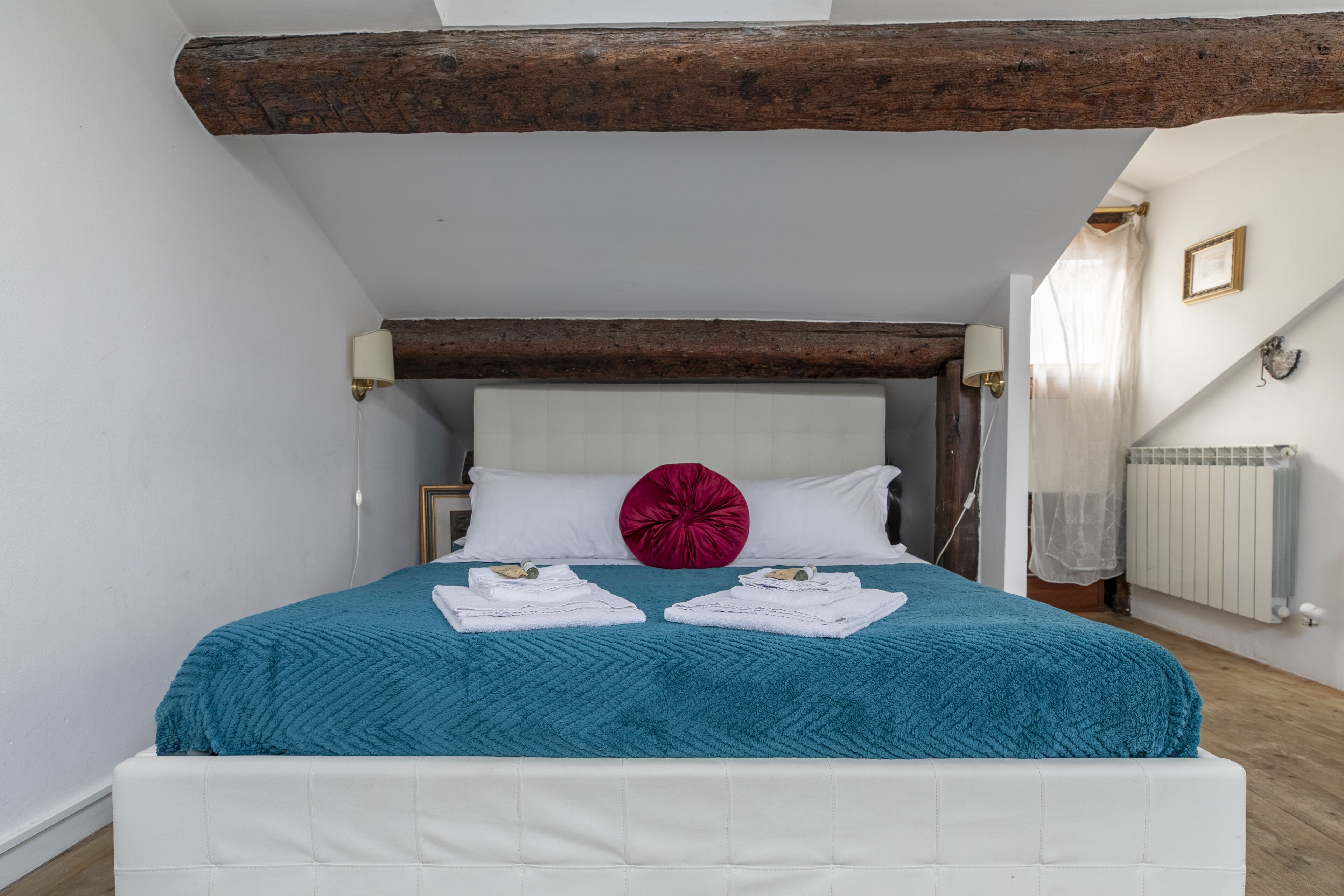 Une chambre avec un lit blanc, une literie de couleur turquoise, un coussin rouge en guise d’élément décoratif, et une poutre en bois au plafond.