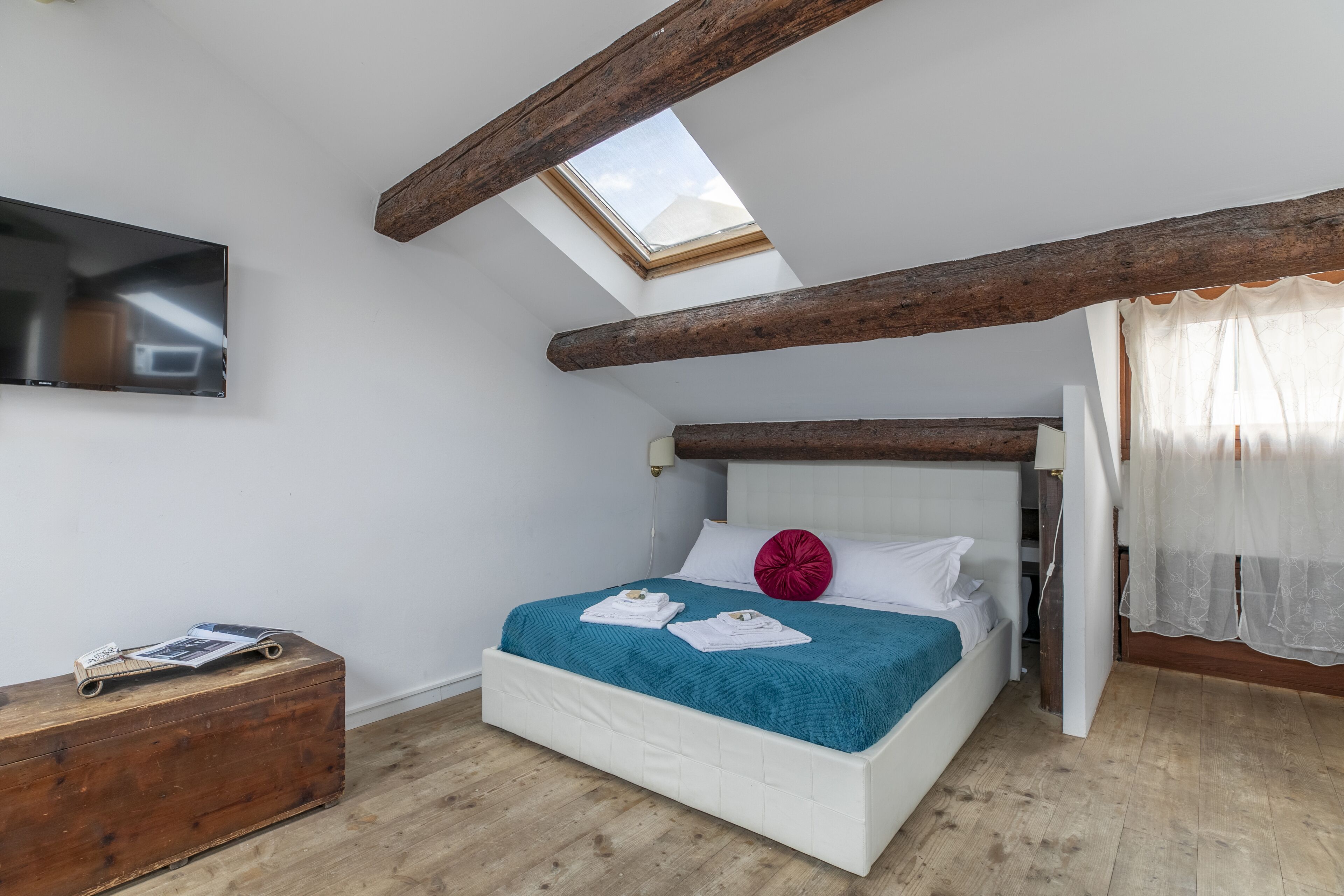 A bedroom with a skylight, a bed with a teal blanket, white pillows, and a wooden chest of drawers.